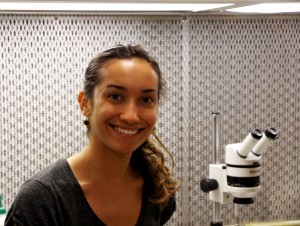 Photo of Camille Prejean smiling in the lab in front of the laminar flow hood