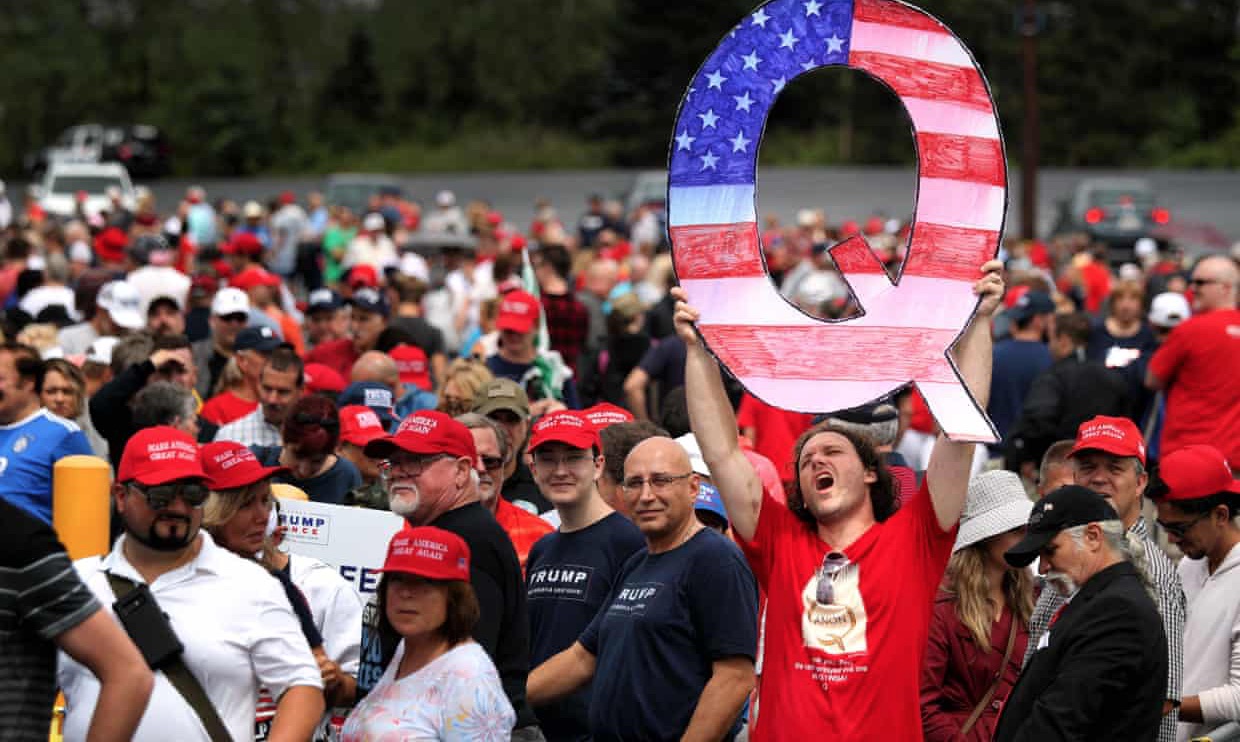 a photo of a man at a political rally holding a large cardboard cutout of a "Q," representing his support for the qanon conspiracy