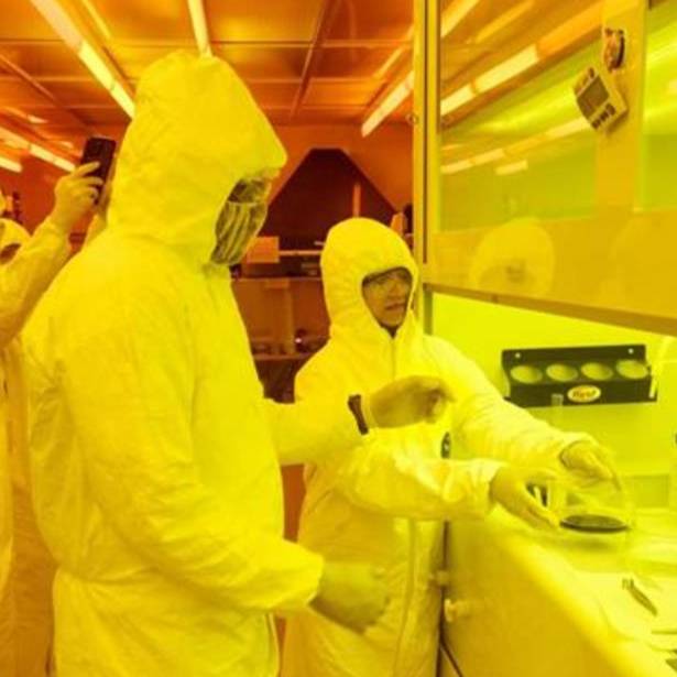 A group of researchers wearing full cleanroom suits and protective eyewear work under yellow lighting inside a lab. One person appears to be handling a sample at a fume hood while others observe and document the process.