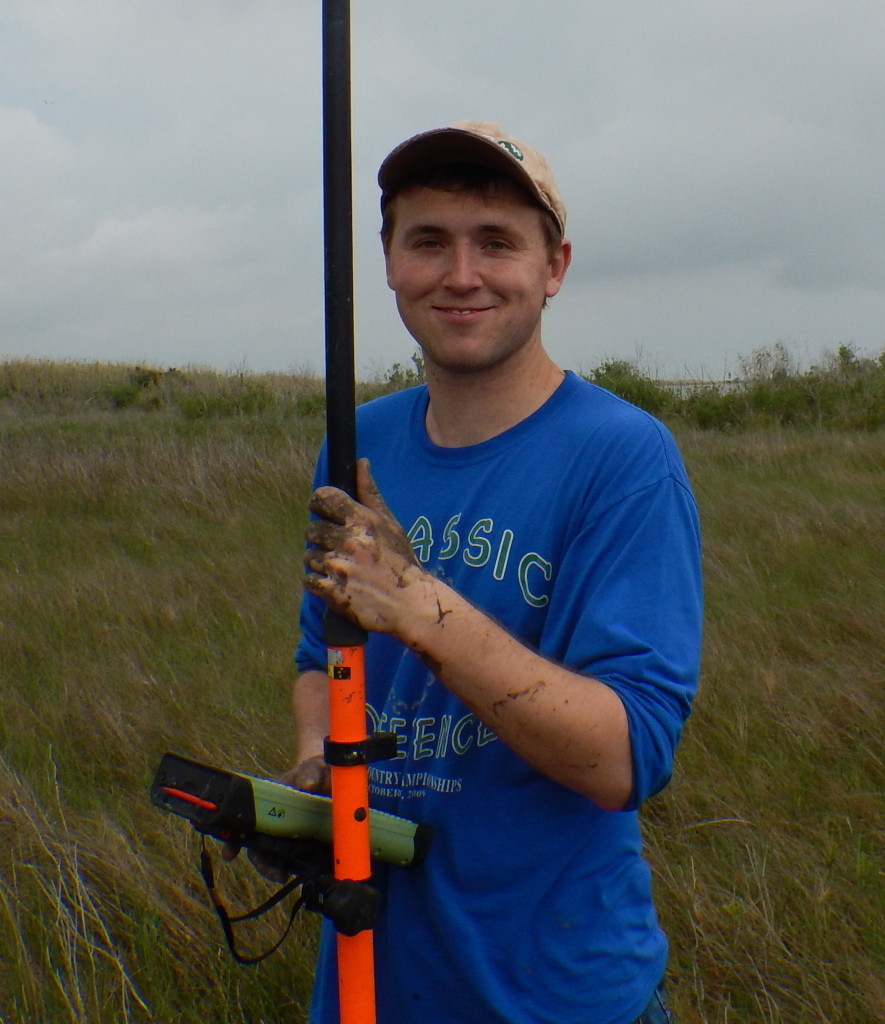 Man stands in a marsh holding a long orange pole connected to a handheld computer.