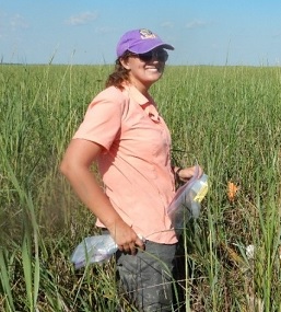 Woman with baseball cap stands in grassland, smiling and holding plastic bags.