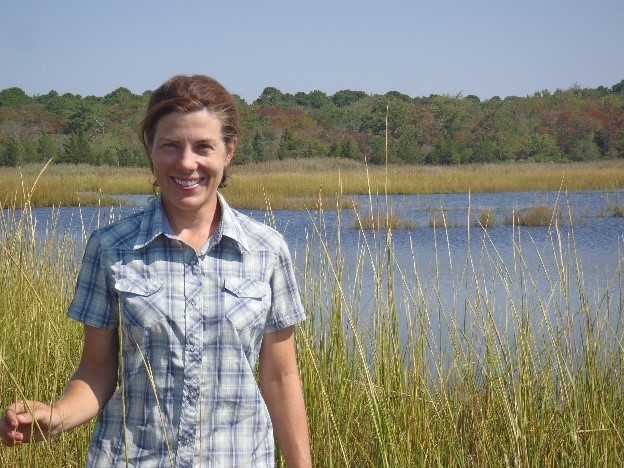 Woman stands in a wetland with lots of grass, water, and trees in the background.